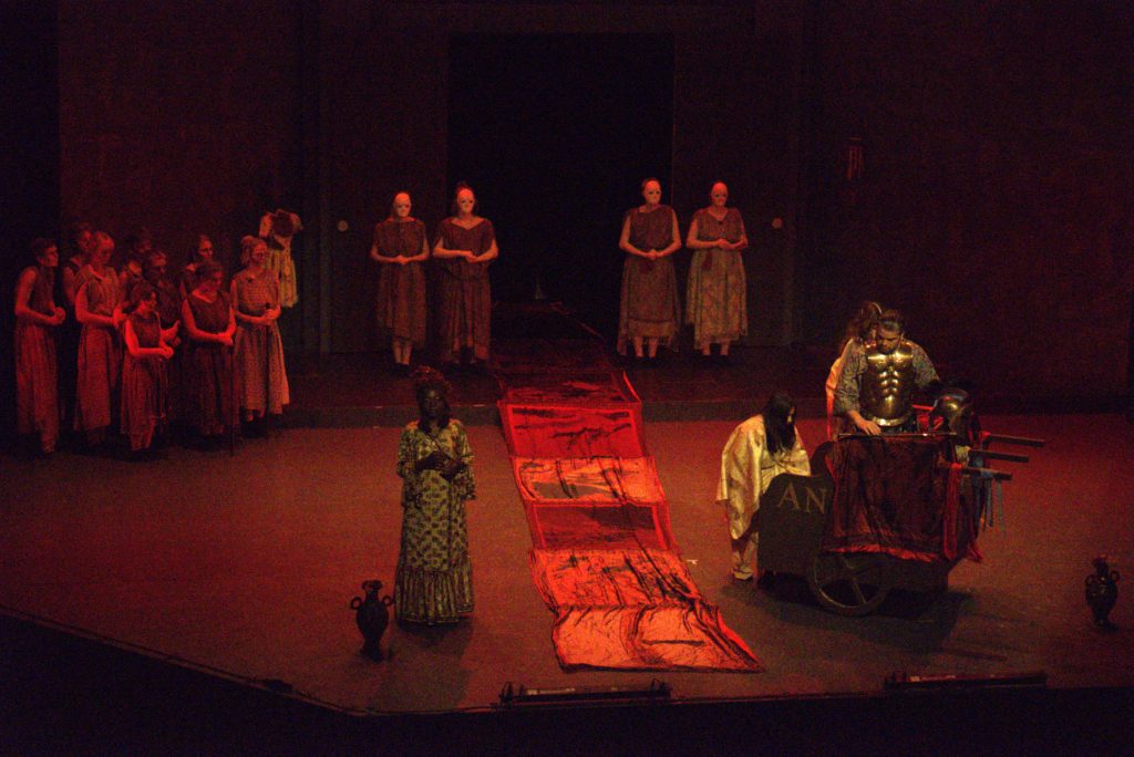 The tapestry scene, the furies stand by the doors of the house, Clytemnestra addresses Agamemnon, who is still in the chariot with Cassandra, while chorus of elders watch
